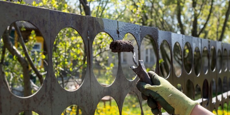 a hand with a paint roller paints an iron fence with brown paint in the garden